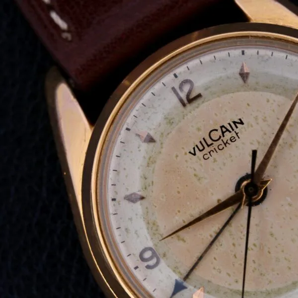Close-up of a Vulcain Cricket 17 Jewels wristwatch with a gold-toned case, weathered cream dial featuring numbered hour markers, and pointed hands. Partially visible is its brown leather strap.