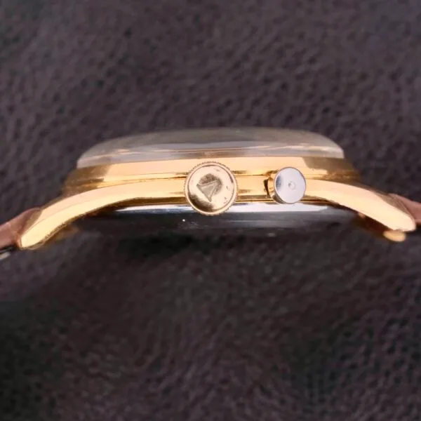A side view of the Vulcain Cricket 17 Jewels wristwatch highlights its gold casing and round crown on a textured dark surface. The watch features a brown leather strap and a clear, slightly domed glass face, completing its elegant design.