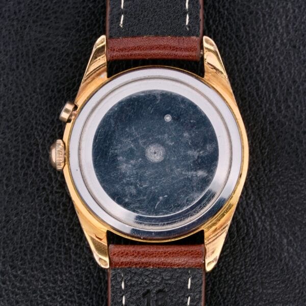 Back view of a Vulcain Cricket 17 Jewels wristwatch with a gold-toned case and brown leather strap on a black textured surface, showcasing its intricate mechanism without cover.