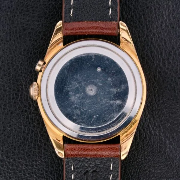 Back view of a Vulcain Cricket 17 Jewels wristwatch with a gold-toned case and brown leather strap on a black textured surface, showcasing its intricate mechanism without cover.