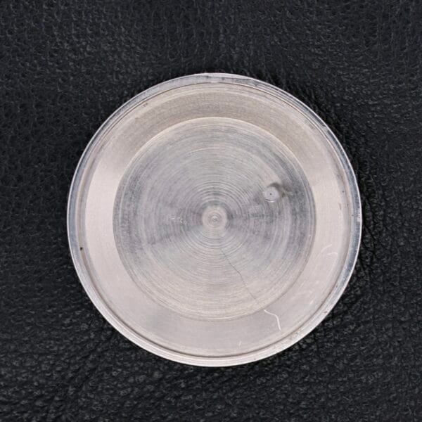 A close-up of a round, transparent object with visible texturing resembling the intricate detailing of the Vulcain Cricket 17 Jewels, featuring a small bump on its surface against a black, textured background.