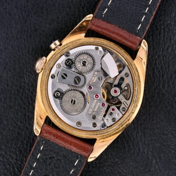Close-up of the Vulcain Cricket 17 Jewels mechanical watch with a transparent back, showcasing its intricate gears and components. The gold case pairs beautifully with a brown and black leather strap, elegantly resting on a black leather surface.