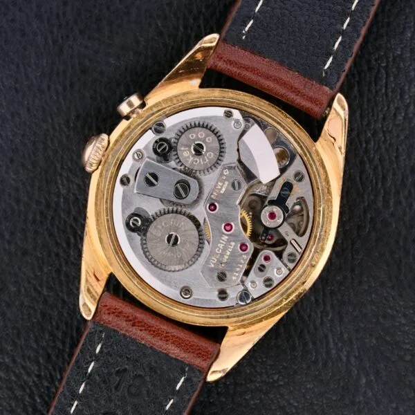 Close-up of the Vulcain Cricket 17 Jewels mechanical watch with a transparent back, showcasing its intricate gears and components. The gold case pairs beautifully with a brown and black leather strap, elegantly resting on a black leather surface.