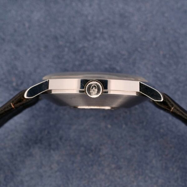 Side view of a sleek, silver Movado Sub-Sea Tempomatic Date wristwatch with a black leather strap on a textured blue surface. The prominently visible crown highlights the watchs minimalist design.