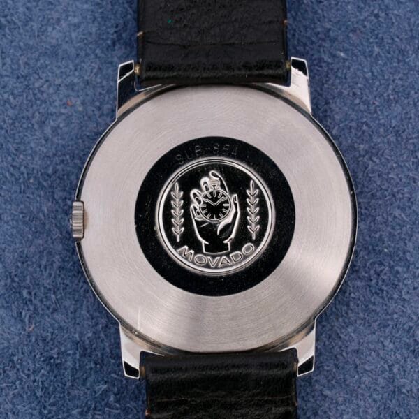 The back of the silver Movado Sub-Sea Tempomatic Date watch features an engraved logo with a hand holding a tool, surrounded by leaves, while Sub-Sea, Movado, and Tempomatic Date elegantly adorn the blue textured background. The watch has a black leather strap.