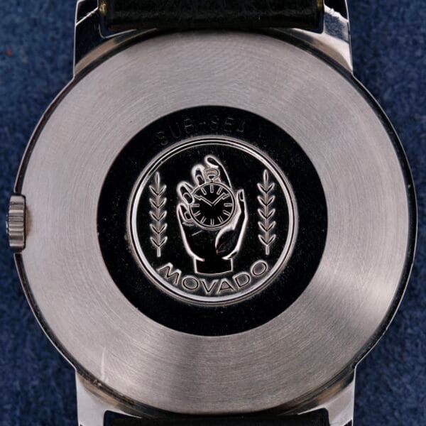 The back of the silver Movado Sub-Sea Tempomatic Date watch shows an engraved hand holding a watch with laurel leaves. MOVADO is engraved below, while Tempomatic Date is etched above, and it features a black strap.