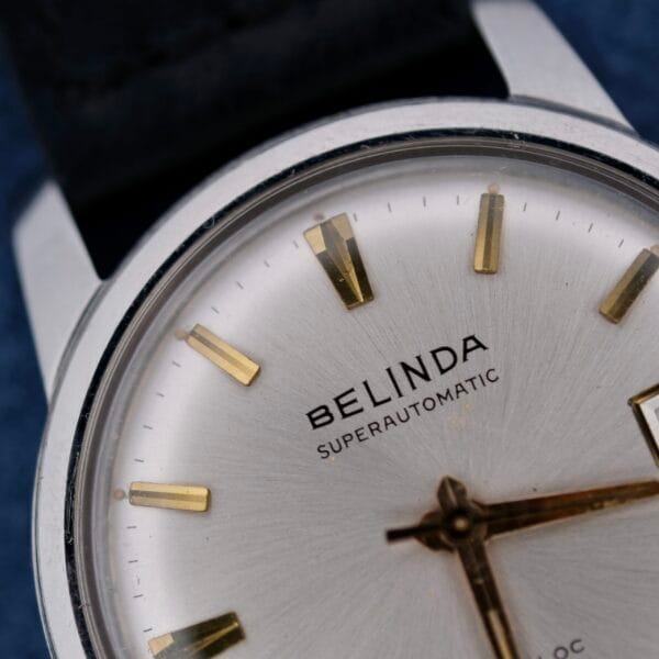 Close-up of a wristwatch with a silver face, gold hour markers and hands, and the name Belinda Super Automatic Date elegantly printed on the dial. It features a sleek metallic case and what appears to be a genuine leather strap.