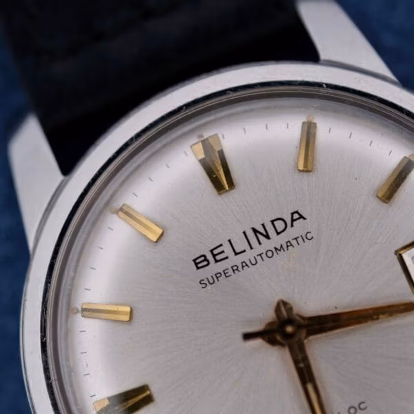 Close-up of a wristwatch with a silver face, gold hour markers and hands, and the name Belinda Super Automatic Date elegantly printed on the dial. It features a sleek metallic case and what appears to be a genuine leather strap.