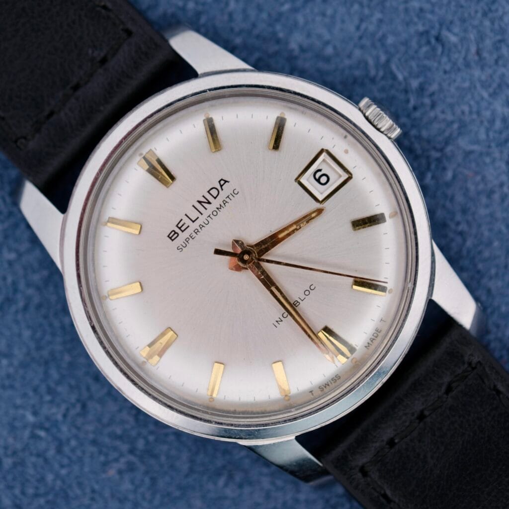A close-up of a vintage Belinda Super Automatic Date wristwatch showcases its silver dial, gold hour markers, and date display reading 6. It rests on a blue textured surface and features a classic black leather strap.