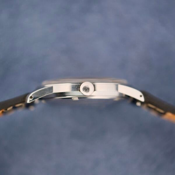Close-up side view of the elegant Belinda Super Automatic Date wristwatch against a soft blue background. The watch boasts a thin profile and prominent crown, with a sleek silver case beautifully complementing the partially visible black strap.