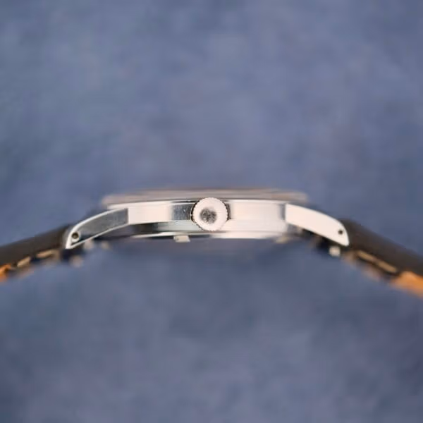 Close-up side view of the elegant Belinda Super Automatic Date wristwatch against a soft blue background. The watch boasts a thin profile and prominent crown, with a sleek silver case beautifully complementing the partially visible black strap.