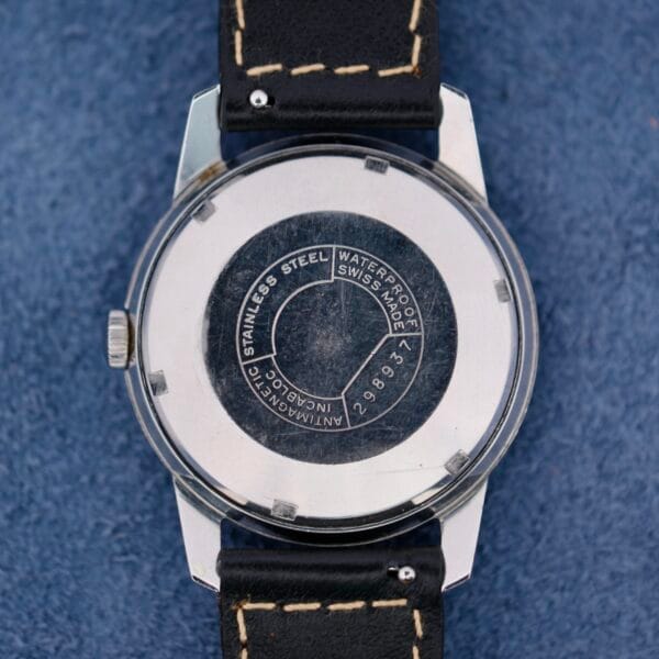 The back of a Belinda Super Automatic Date wristwatch with a stainless steel case and black leather strap displays Waterproof, Swiss Made, and other specifications on a textured blue surface.