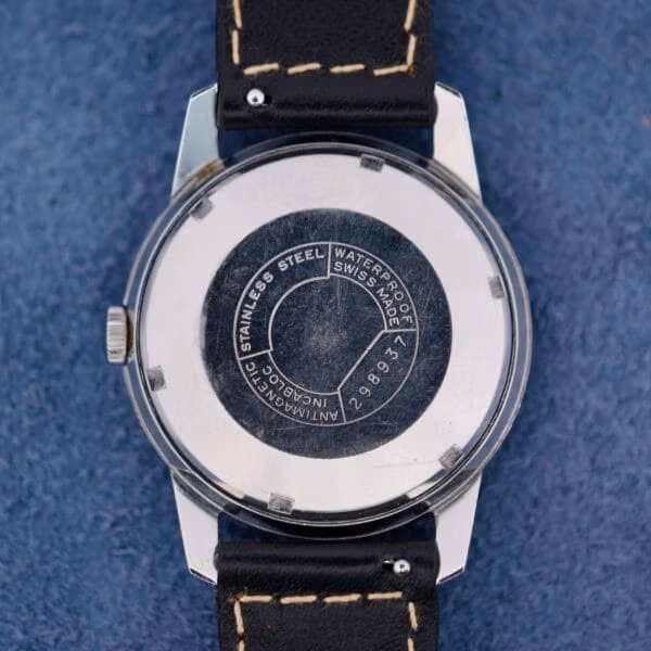 The back of a Belinda Super Automatic Date wristwatch with a stainless steel case and black leather strap displays Waterproof, Swiss Made, and other specifications on a textured blue surface.