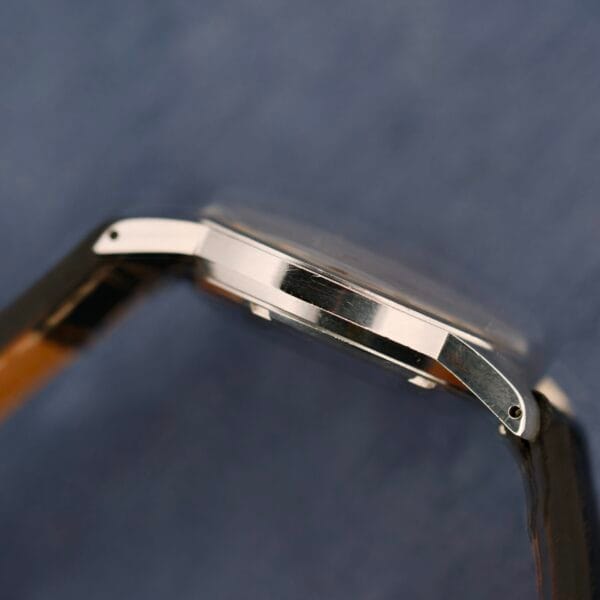Close-up of a Belinda Super Automatic Date wristwatch, featuring a smooth metallic side profile, silver finish, and black leather strap against a blue textured background.