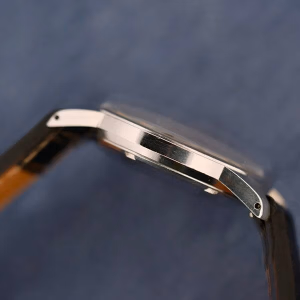 Close-up of a Belinda Super Automatic Date wristwatch, featuring a smooth metallic side profile, silver finish, and black leather strap against a blue textured background.