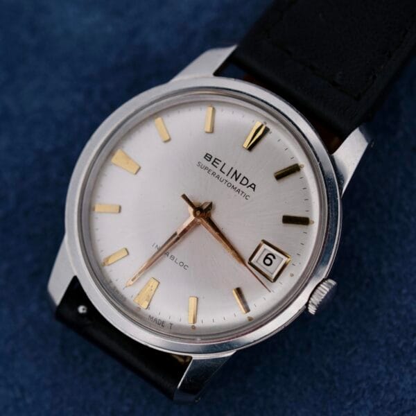 A close-up of the Belinda Super Automatic Date wristwatch shows a silver face with gold hour markers, displaying the 6th. The sleek stainless steel case is complemented by a black leather strap, exuding elegance.