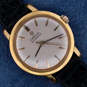 Close-up of the Omega Automatic Ladies wristwatch in gold with a black leather strap, displayed on a blue surface.