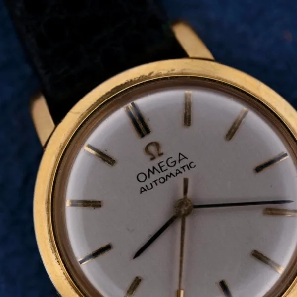 A close-up of the Omega Automatic Ladies wristwatch, showcasing a gold case, white dial with simple hour markers, and black leather strap against a striking blue background.