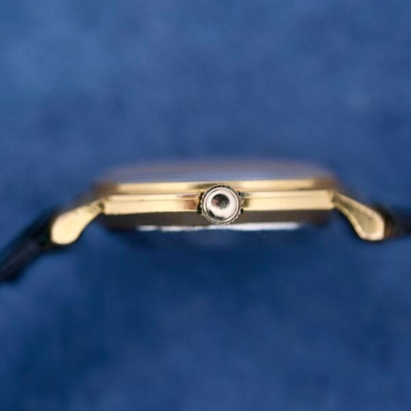 A gold Omega Automatic Ladies wristwatch is shown close-up against a blurred blue background, with focus on the crown to highlight its metallic structure and details.