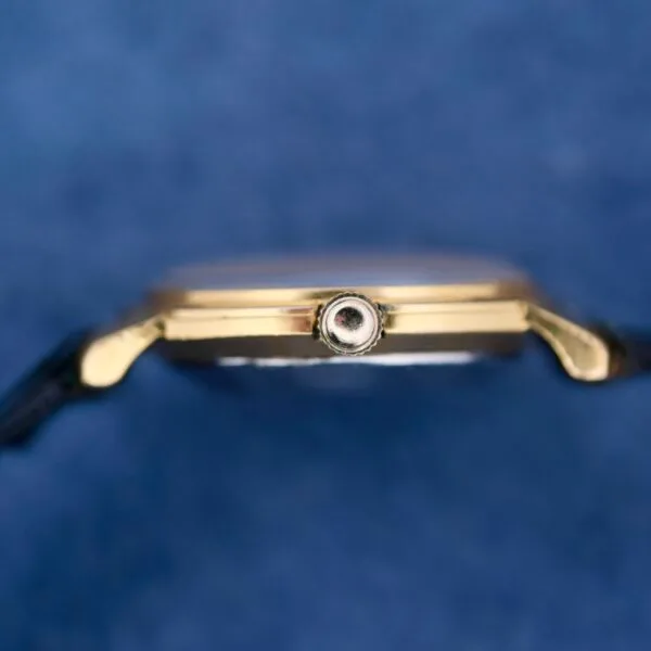 A gold Omega Automatic Ladies wristwatch is shown close-up against a blurred blue background, with focus on the crown to highlight its metallic structure and details.