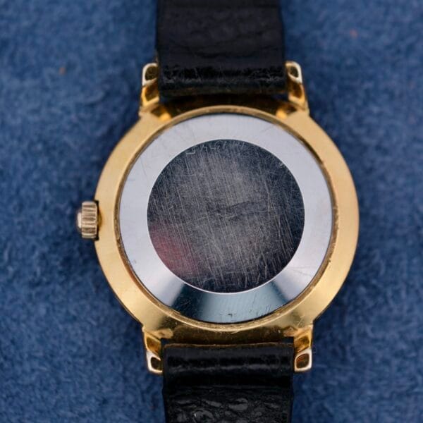 The image shows the scratched back of a vintage Omega Automatic Ladies wristwatch, featuring a black leather strap and a gold casing, elegantly placed on a blue textured surface.