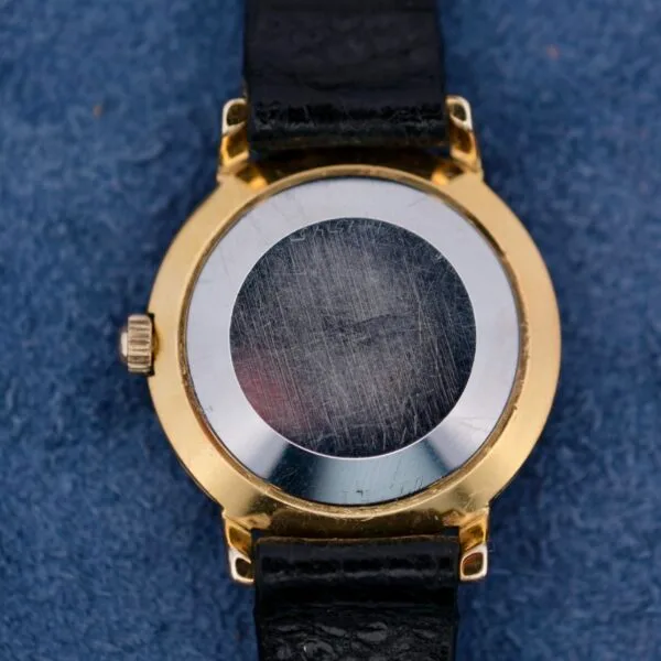The image shows the scratched back of a vintage Omega Automatic Ladies wristwatch, featuring a black leather strap and a gold casing, elegantly placed on a blue textured surface.