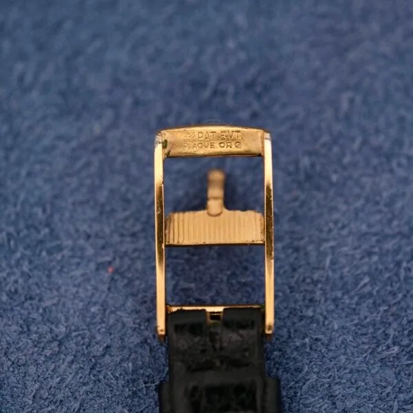 A close-up of a gold watch buckle stamped with Patek Philippe on a textured blue background evokes elegance reminiscent of the Omega Automatic Ladies, with an attached black leather strap visible at the bottom.