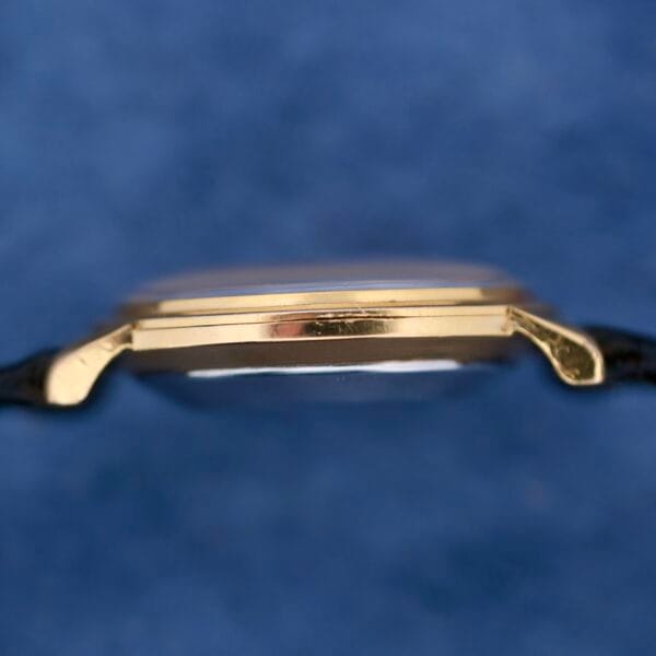 Close-up of the Omega Automatic Ladies wristwatch with a gold slim profile and black strap against a soft blue background.