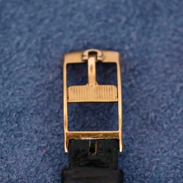 Close-up of a gold rectangular watch buckle with a textured bar on a black leather strap, evoking the Omega Automatic Ladies design, set against a blue textured background.