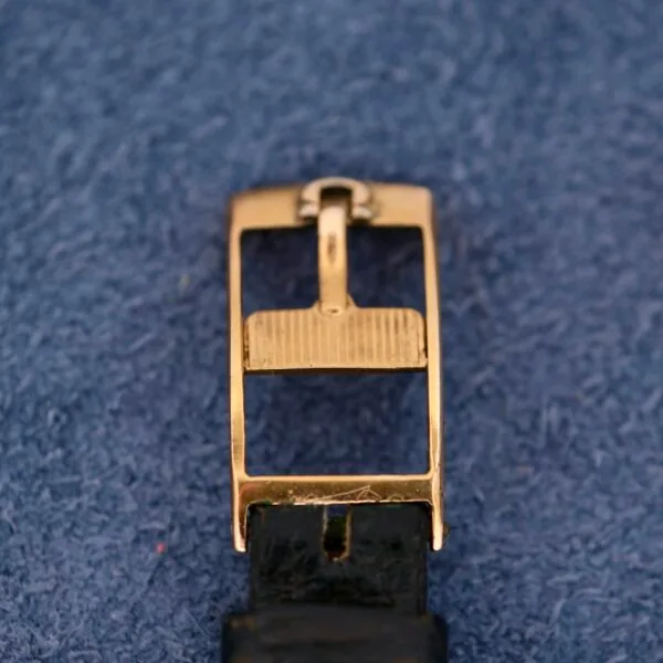 Close-up of a gold rectangular watch buckle with a textured bar on a black leather strap, evoking the Omega Automatic Ladies design, set against a blue textured background.