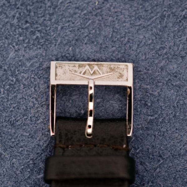 A close-up of a silver belt buckle with a geometric design, evoking the style of a Movado Sub-Sea Tempomatic Date, attached to a black leather belt on a textured blue background.