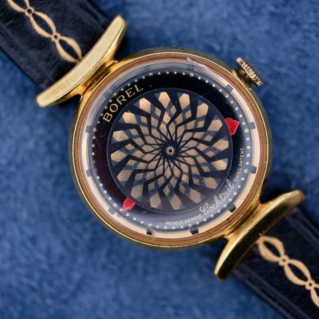 The Ernest Borel Kaleidoscope Cocktail watch features a spinning kaleidoscope dial and a black leather strap, displayed on a blue background.