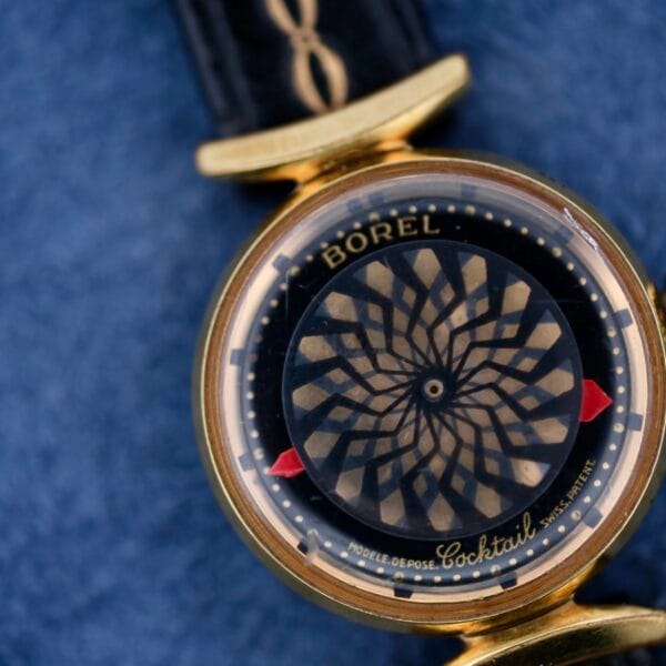 The Ernest Borel Kaleidoscope Cocktail watch, with a vintage hypnotic swirling dial, features a gold case and black leather strap set against a textured blue background.