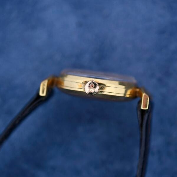 Side view of an Ernest Borel Kaleidoscope Cocktail gold wristwatch with the winding crown, set against a textured blue background. It features a sleek black band and exudes kaleidoscope elegance.