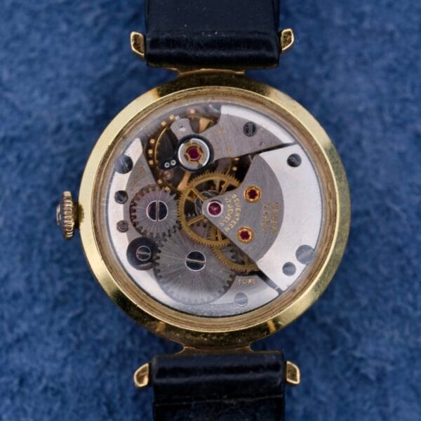 A close-up of the Ernest Borel Kaleidoscope Cocktails mechanical watch movement reveals intricate gears and cogs. The timepiece boasts a gold outer case, a black leather strap, set against a textured dark blue fabric backdrop.