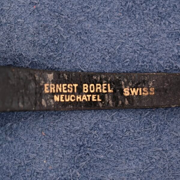 A close-up of a black strap on a blue textured background highlights the elegant craftsmanship, embossed with Ernest Borel Neuchatel Swiss, reflecting the artistry of the Ernest Borel Kaleidoscope Cocktail.