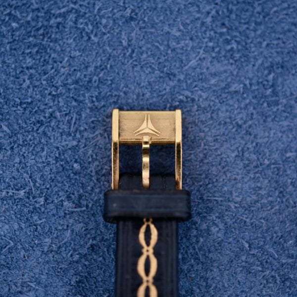 Close-up of a black belt with gold accents on a textured blue surface, echoing the intricate beauty of the Ernest Borel Kaleidoscope Cocktail. The gold buckle has an engraved emblem, adding elegance to the accessory.