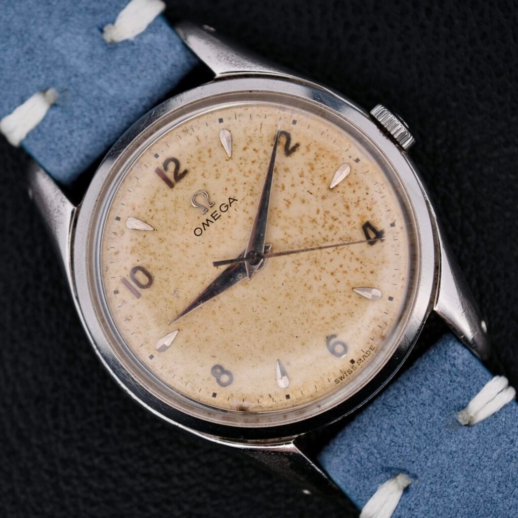 Omega Pre Seamaster Cal. 283 wristwatch with a speckled cream dial and blue leather strap, showcased on a black background.