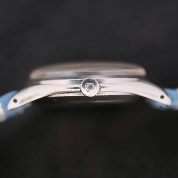 A side view close-up of an Omega Pre Seamaster Cal. 283 watch highlights the engraved logo on its crown. The metal case and light-colored strap stand out against a dark background, creating a striking contrast.