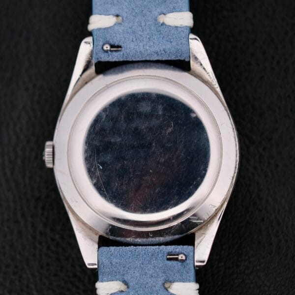 A close-up of the back of an Omega Pre Seamaster Cal. 283 wristwatch, showcasing its silver casing and light blue suede strap against a black textured background, emphasizing its elegance and precision.
