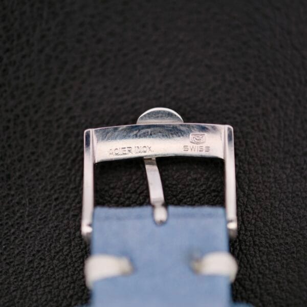 A close-up of a silver watch buckle engraved with ACIER INOX and SWISS evokes the Omega Pre Seamaster Cal. 283 legacy. The buckle is attached to a light blue strap against a textured dark background.