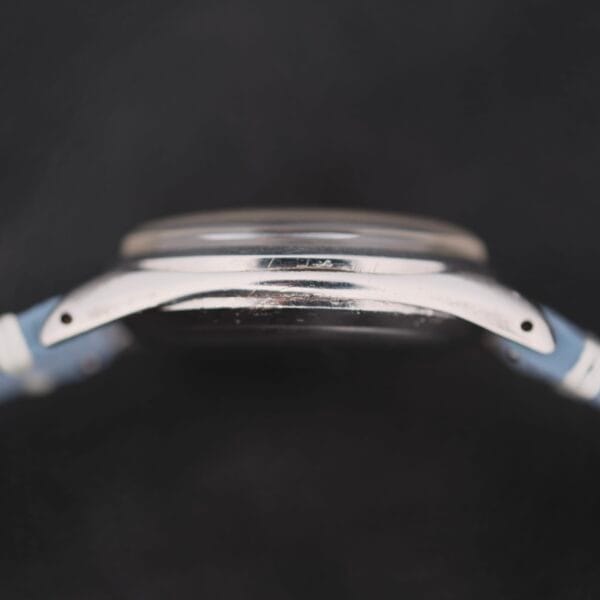 Close-up side view of an Omega Pre Seamaster Cal. 283 with a round metallic case and light blue strap, its face slightly obscured to emphasize the sleek, minimalist design against a dark background.