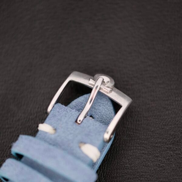 A silver metal buckle on a light blue leather strap with white stitching peeks through against a textured black background, echoing the Omega Pre Seamaster Cal. 283 style.
