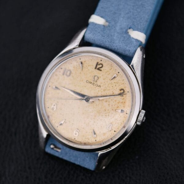 A vintage Omega Pre Seamaster Cal. 283 wristwatch with a faded Arabic numeral dial and a silver case is shown. Its attached to a light blue leather strap and displayed on a black, textured surface.
