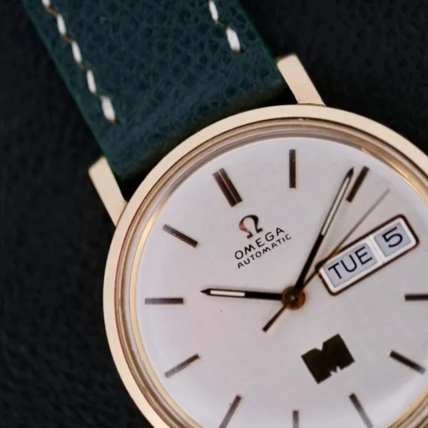 Close-up of an Omega Automatic Day/Date Linen M Dial watch featuring a gold-tone case and sleek black leather strap. The dial displays the day/date: Tuesday, the 5th, graced by the iconic Omega logo with understated black hour markings and hands.