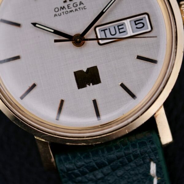 Close-up of an Omega Automatic Day/Date Linen M Dial watch featuring gold casing, white dial with black hour markers and hands, displaying TUE 5, and a green textured leather strap.
