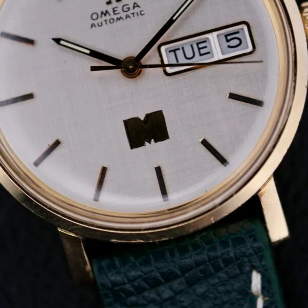Close-up of an Omega Automatic Day/Date Linen M Dial watch featuring gold casing, white dial with black hour markers and hands, displaying TUE 5, and a green textured leather strap.