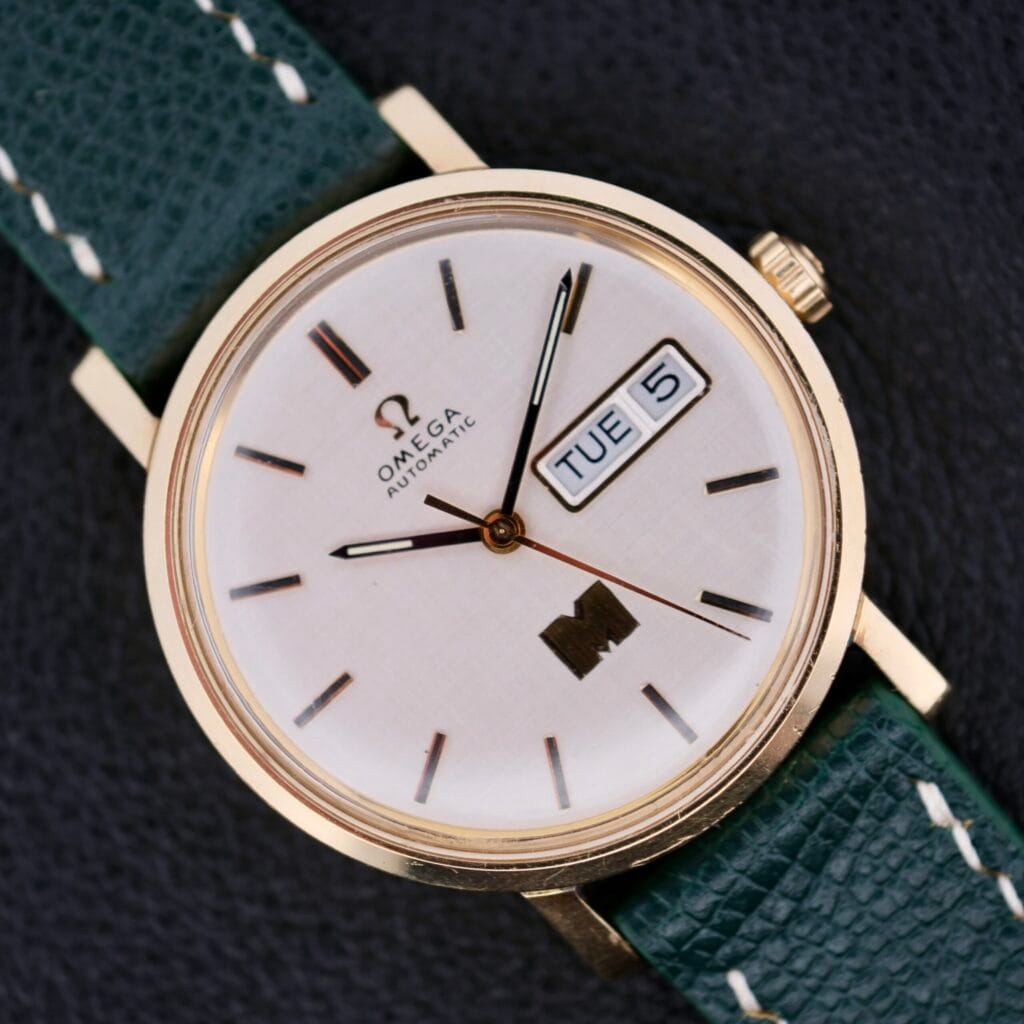 Omega Automatic Day/Date Linen M Dial wristwatch featuring a green leather strap, white linen dial, and day-date display set to Tuesday the 5th.