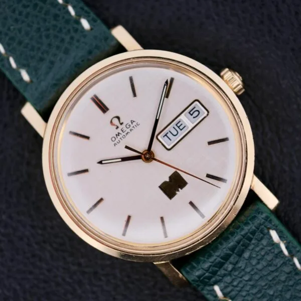 A close-up of the Omega Automatic Day/Date Linen M Dial watch, featuring a gold case and green leather strap. The elegant Linen M Dial displays the time, Tuesday, and 5th against a textured dark background.