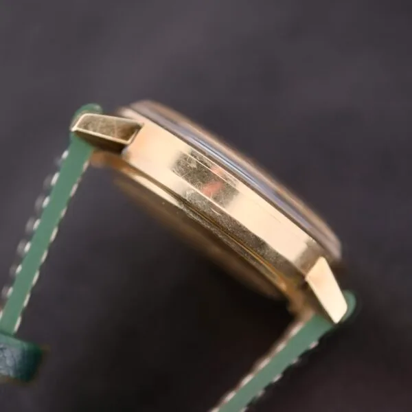 Close-up of an Omega Automatic Day/Date Linen M Dial wristwatch, showcasing a gold-finished case with visible side scratches and green stitched leather straps that enhance its timeless charm.